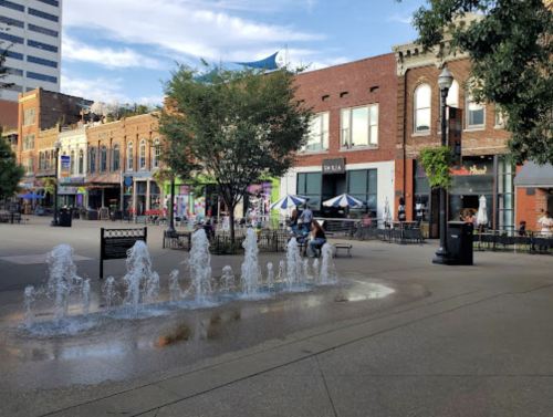A city square with a fountain, benches, and shops, featuring people enjoying the outdoor space on a sunny day.
