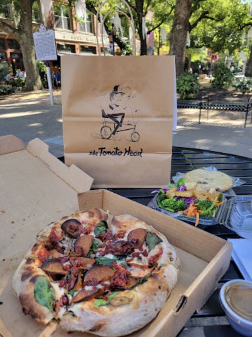 A pizza with toppings, a salad, and a takeout bag from "The Tomito Head" on a table in a park.