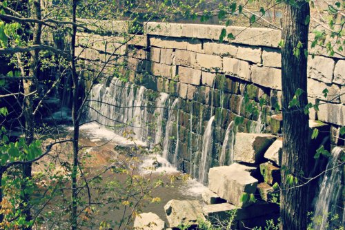 A stone dam with water cascading over it, surrounded by trees and greenery.