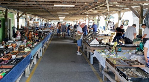 A bustling indoor market with vendors and shoppers browsing various antiques and collectibles along long tables.
