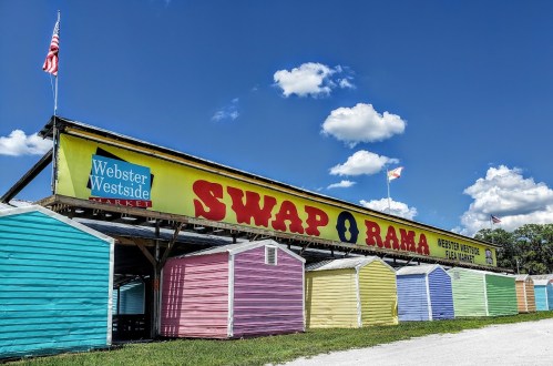 Colorful market stalls under a blue sky with clouds, featuring a large "SWAP-O-RAMA" sign.