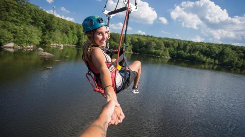 A person zip-lining over a lake, smiling and reaching out to hold another person's hand. Lush greenery in the background.