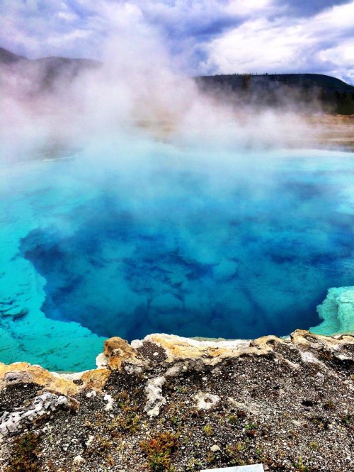 A vibrant blue hot spring surrounded by steam and rocky terrain, set against a cloudy sky.