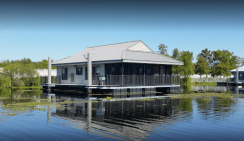 A modern house on stilts surrounded by calm water and greenery, reflecting in the water's surface.