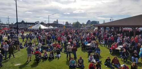 A large crowd gathers at an outdoor event, with tents and a pavilion in the background under a partly cloudy sky.