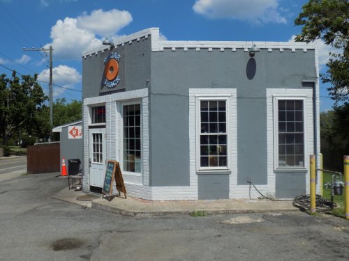 A small gray building with large windows, featuring a round sign, surrounded by trees and a clear blue sky.