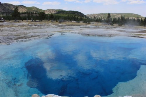 A vibrant blue hot spring surrounded by rocky terrain and trees under a clear sky in a natural landscape.