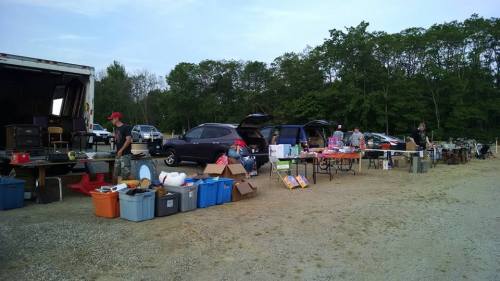 A bustling outdoor market with various items for sale, cars parked nearby, and people browsing through goods.
