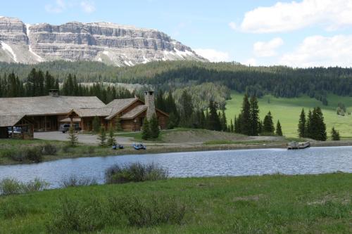 A scenic view of a log cabin by a lake, surrounded by lush greenery and mountains under a partly cloudy sky.