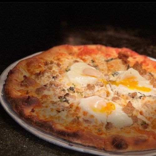 A freshly baked pizza topped with eggs, sausage, and herbs on a rustic wooden table.