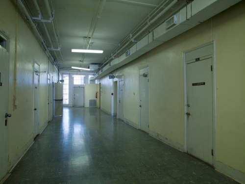 A long, empty hallway with yellow walls and several closed doors, illuminated by overhead lights.