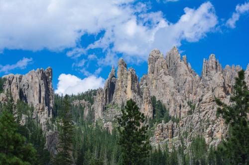 A rugged mountain landscape with towering rock formations and lush green trees under a blue sky with fluffy clouds.