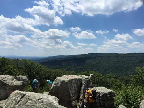 Two hikers explore rocky terrain with a lush green valley and blue sky in the background.