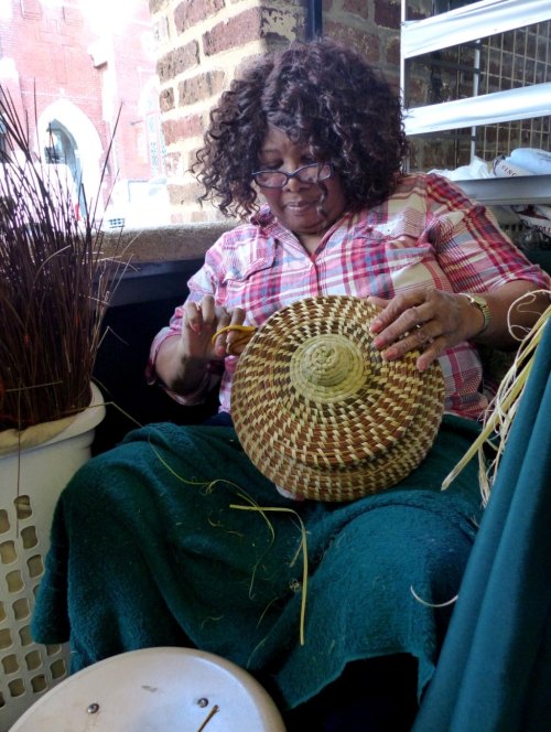 A woman with curly hair weaves a basket, sitting on a chair with a green blanket draped over her lap.