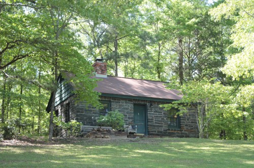 A small stone cabin surrounded by lush green trees and grass, featuring a chimney and a simple front door.