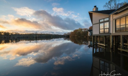 A serene lake at sunset, reflecting clouds and trees, with a modern house on the shore.