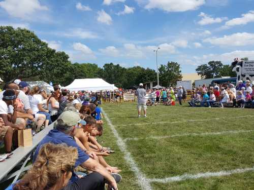 A crowd watches a sporting event on a sunny day, with participants and tents in the background on a grassy field.