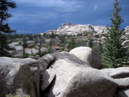 Rocky landscape with large boulders and pine trees under a dramatic, cloudy sky.