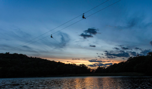 Two zipliners glide over a lake at sunset, with a colorful sky and silhouettes of trees in the background.