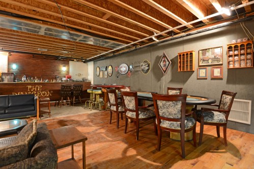 A cozy basement bar with wooden floors, a bar counter, and tables surrounded by chairs, featuring vintage decor on the walls.