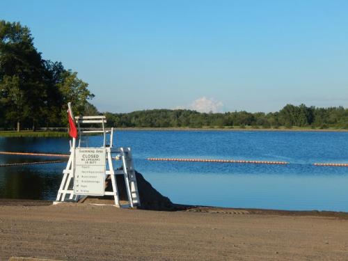 A lifeguard stand by a closed swimming area at a calm lake, with a sign indicating closure and a red flag.