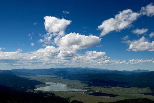 A panoramic view of a valley with a lake, surrounded by mountains and a bright blue sky filled with fluffy clouds.