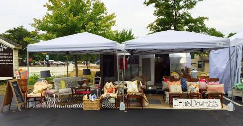 A market stall with two tents displaying various furniture, pillows, and decor items, set up outdoors.