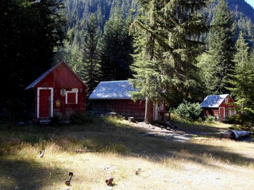 Two rustic red cabins surrounded by tall trees and a grassy area, set against a backdrop of mountains.