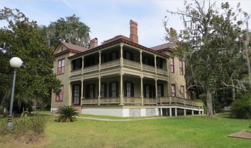 A large, historic two-story house with a wraparound porch, surrounded by greenery and trees draped in Spanish moss.
