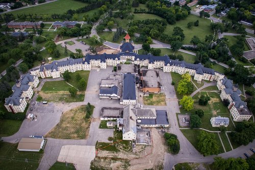 Aerial view of a large, abandoned building complex surrounded by greenery and roads.