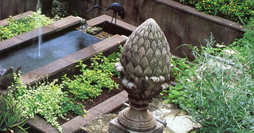 A stone artichoke sculpture stands in a lush garden with a pond and greenery, featuring a decorative bird in the background.