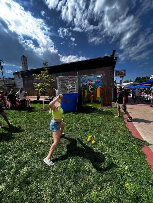 A girl in a yellow top throws a ball at a colorful carnival game, with people and tents in the background.