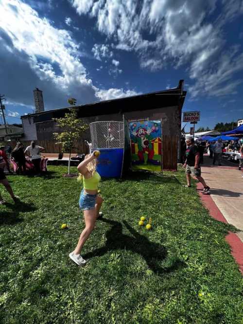 A girl in a yellow top throws a ball at a colorful carnival game, with people and tents in the background.