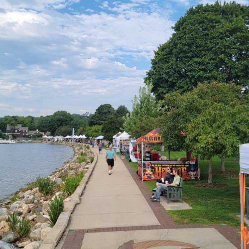 A scenic lakeside path with vendors and tents, surrounded by greenery and a cloudy sky. People stroll along the walkway.