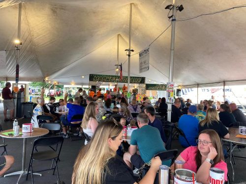 A crowded tent filled with people dining and socializing at a market event, with colorful stalls in the background.