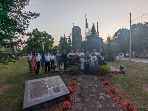 A group of people in traditional attire gathered at a memorial site with flags and flowers, surrounded by trees.