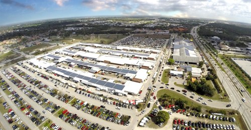Aerial view of a large parking lot with numerous cars and buildings, surrounded by green areas and roads.