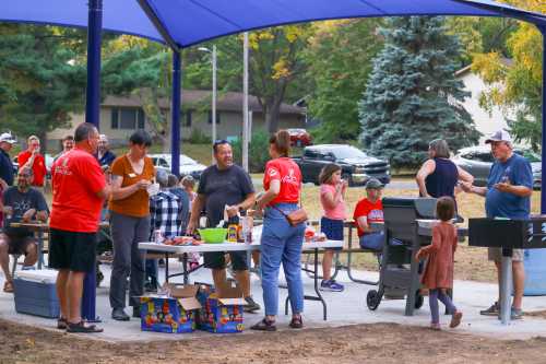 A community gathering at a park with people enjoying food, chatting, and grilling under a large blue canopy.