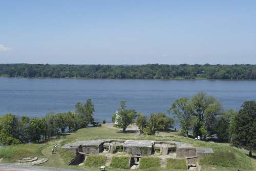 A scenic view of a river with green trees and a historic structure in the foreground under a clear blue sky.