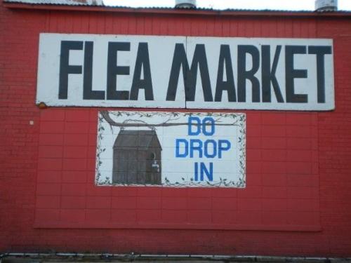 Sign on a red wall reading "Flea Market" with "DO DROP IN" in blue below a graphic of a building.