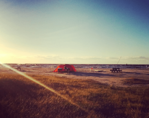 A red tent on a beach at sunset, with picnic tables and gentle waves in the background.