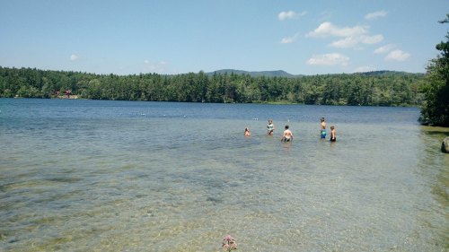 Children playing in shallow water at a clear lake, surrounded by trees and mountains under a blue sky.