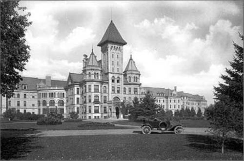 Historic black-and-white photo of a large, ornate building with a tower, surrounded by trees and a vintage car in the foreground.