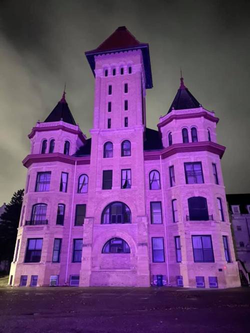 A large, historic building with towers, illuminated in purple light against a dark, cloudy sky.