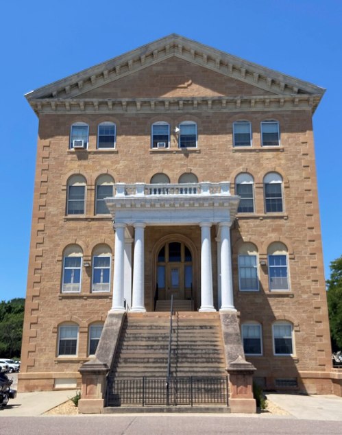 Historic building with a grand entrance, featuring columns and a staircase, set against a clear blue sky.