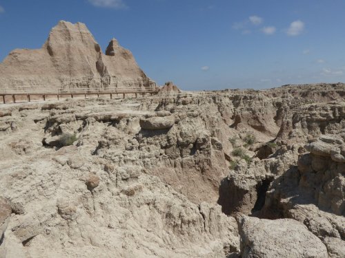 A rugged landscape of eroded rock formations under a blue sky, with a distant peak and scattered vegetation.