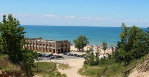A scenic view of a beach with a historic building, trees, and the blue lake in the background under a clear sky.