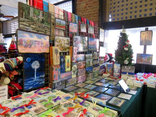 A display of colorful paintings and crafts at a market, featuring various artworks and a small decorated Christmas tree.