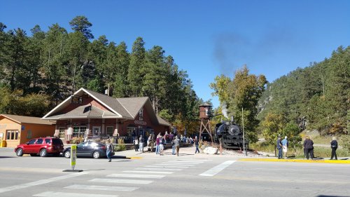 A historic train station with a steam locomotive, surrounded by trees and a crowd of people on a sunny day.