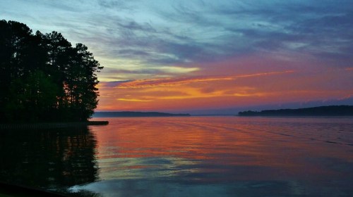 A serene lake at sunset, with vibrant orange and purple skies reflecting on the water, framed by trees on the shore.